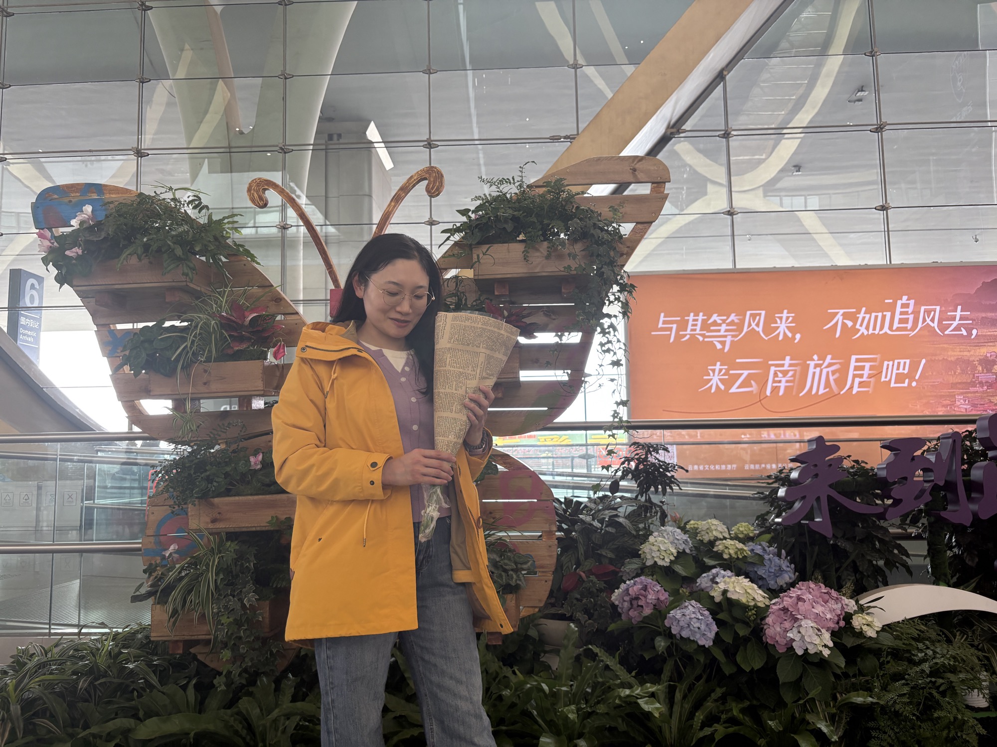 昆明长水机场，晶晶抱着报纸包的花束，站在蝴蝶花艺装置前