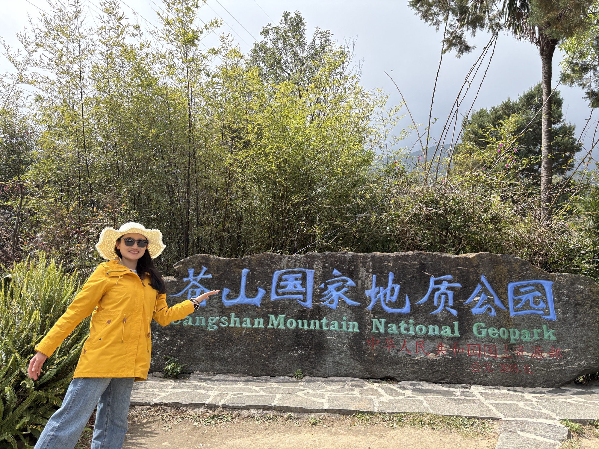 晶晶穿黄色外套站在苍山国家地质公园石碑前，笑着指向石碑